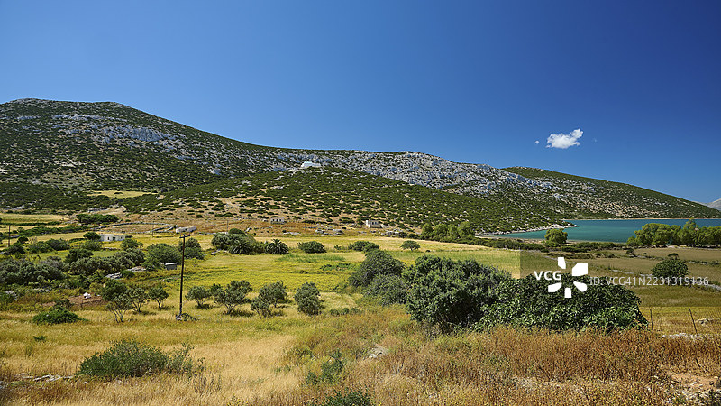 在清澈蓝天下，广阔的黄绿色田野前方是一系列山丘，位于希腊多德卡尼斯群岛的瓦伊湾，阿斯提帕利亚岛。图片素材