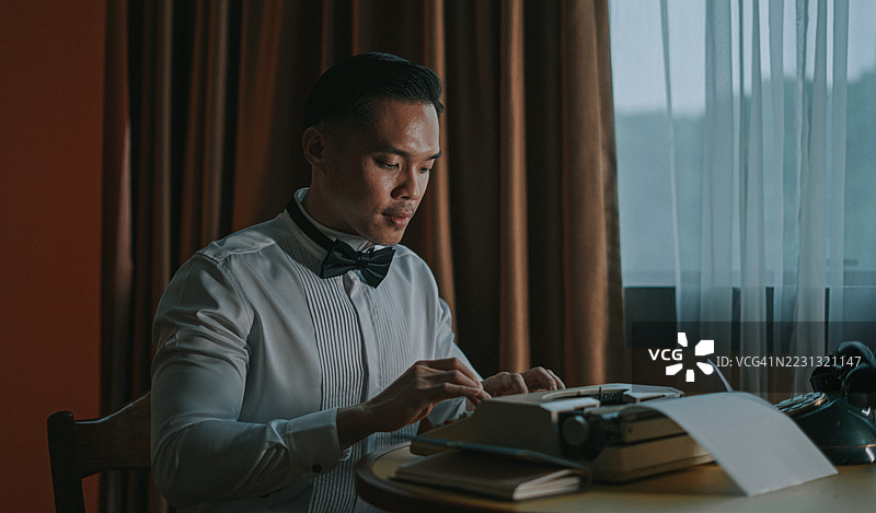年轻亚洲男子穿着正式服装在窗边使用复古打字机打字图片素材