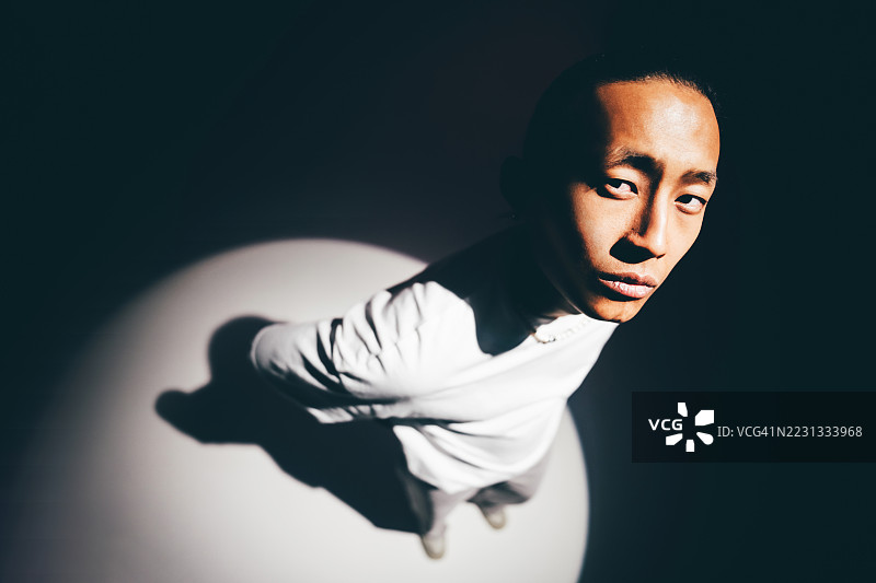 一位亚洲男子在明亮的聚光灯下高角度肖像，影子在黑暗的地面上延伸。图片素材