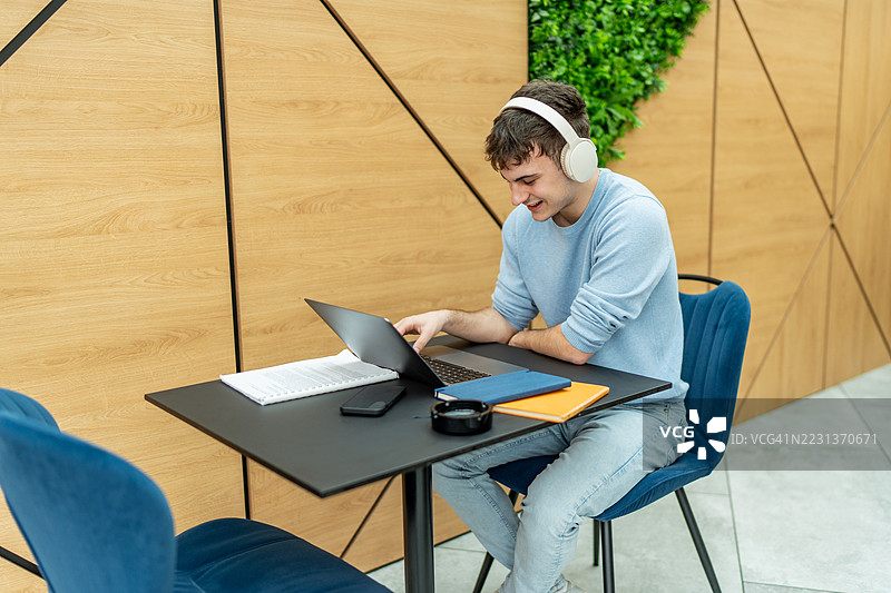 年轻男子在现代共享办公空间中享受在线教育，佩戴着耳机，使用笔记本电脑。图片素材