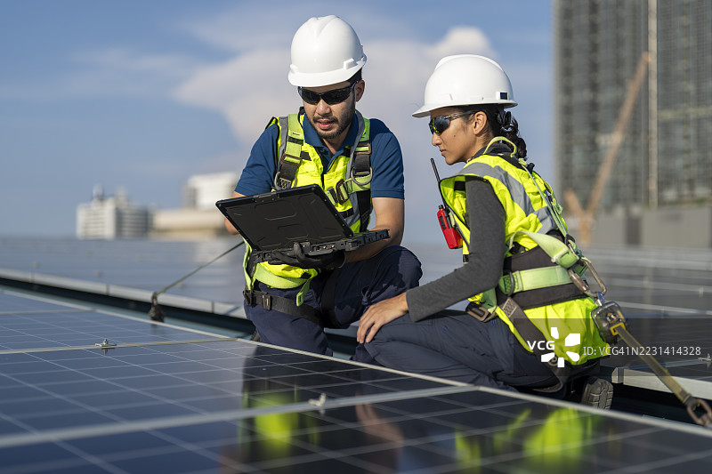 一名男性和一名女性技术人员穿着安全背心和全身安全带，使用笔记本电脑检查建筑屋顶上的太阳能电池板，展现了他们在可再生能源解决方案方面的专业知识。图片素材