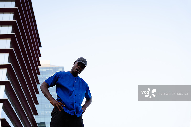 自信的快递员挺直身姿，双手叉腰，城市天际线作为背景，突显其专业风范，身处摩天大楼附近。图片素材