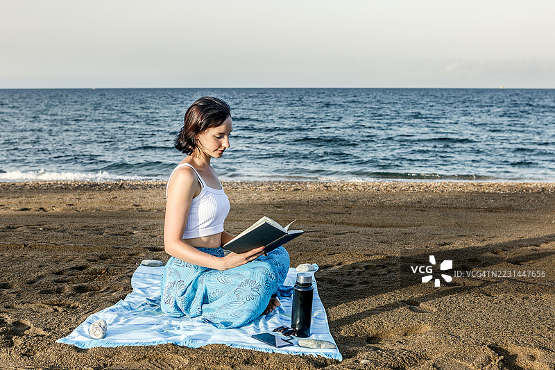 女人在海滩毛巾上阅读书籍，背景是日落时分的海洋图片素材