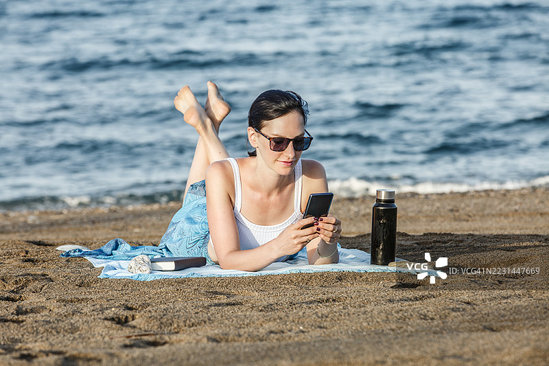女人在海滩上放松，手里拿着智能手机和水瓶图片素材