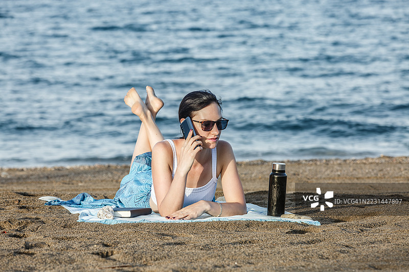 女人在沙滩上放松，打电话，身边是水边的沙滩。图片素材