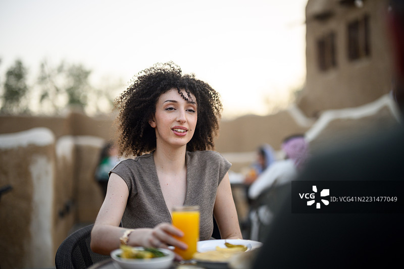 在利雅得一家户外咖啡馆的桌旁，一位卷发年轻女子在与朋友交谈时手中拿着一杯饮料图片素材