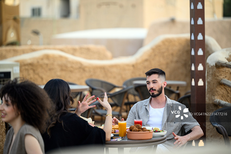 一群年轻的朋友在户外咖啡馆的桌旁聊天，享用饮品，背景是传统的泥砖墙图片素材