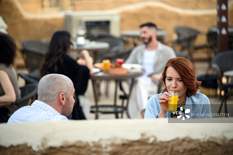 一位成熟的女士在利雅得的户外咖啡馆餐桌旁与朋友交谈时喝橙汁图片素材