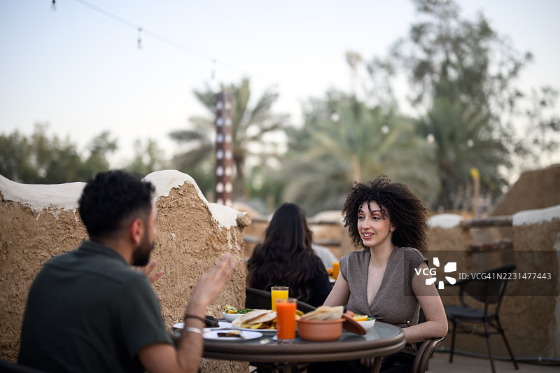 一位卷发女子在沙特古村落的户外咖啡馆桌旁，与一位男士边饮酒边愉快交谈图片素材
