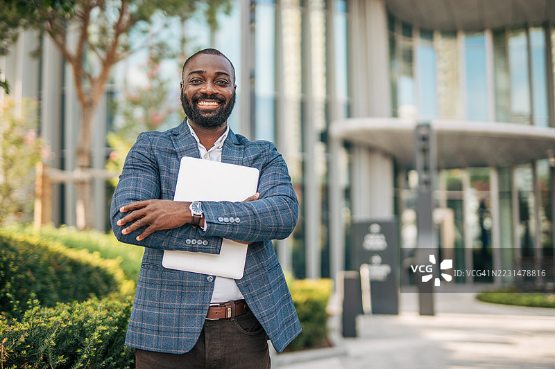 微笑的商人手持笔记本电脑站在办公大楼前图片素材