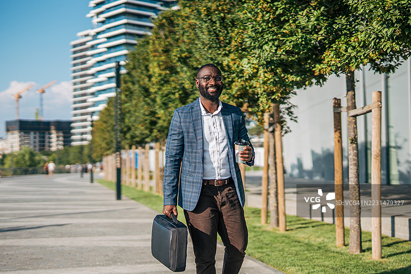 微笑的商人在城市中走着，手里拿着咖啡和公文包图片素材