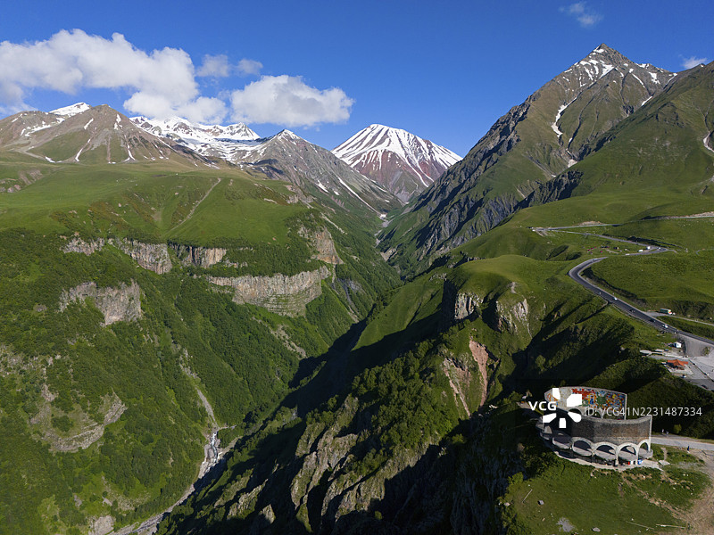 一座圆形建筑上有壁画,坐落在绿色的山坡上,背景是白雪皑皑的山峰,鸟瞰图,格鲁吉亚-俄罗斯友谊纪念碑,观景平台,位于古达乌里和十字山口之间,舍尔霍塔山,姆茨赫塔-姆季安尼地区图片素材