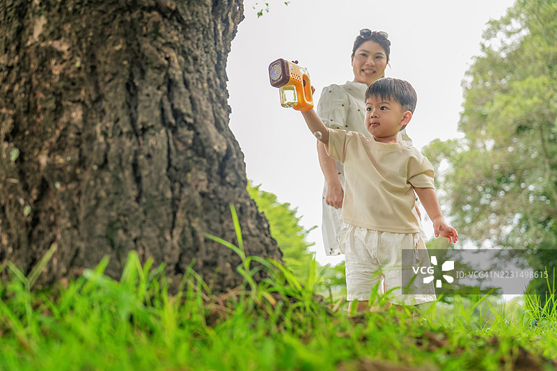 亚洲家庭，母亲和小儿子在户外玩泡泡枪，母子在公园里玩耍，小男孩在空中吹泡泡，母亲微笑着照顾他。图片素材