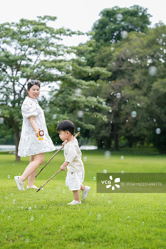 亚洲家庭，母亲和小儿子在户外玩泡泡枪，母子在公园里玩耍，小男孩在空中吹泡泡，母亲微笑着照顾他。图片素材