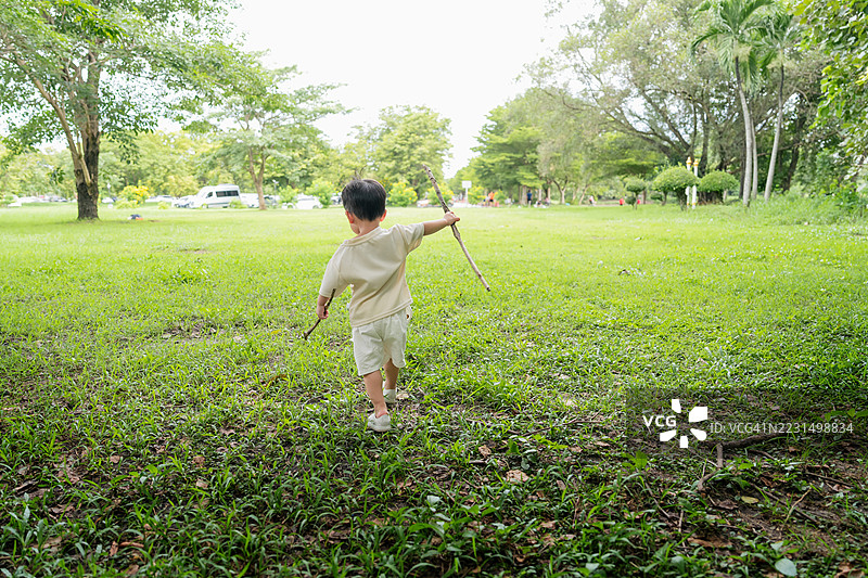 小男孩在阳光明媚的公园里快乐地奔跑，手里拿着木棍，享受探索自然的乐趣。图片素材