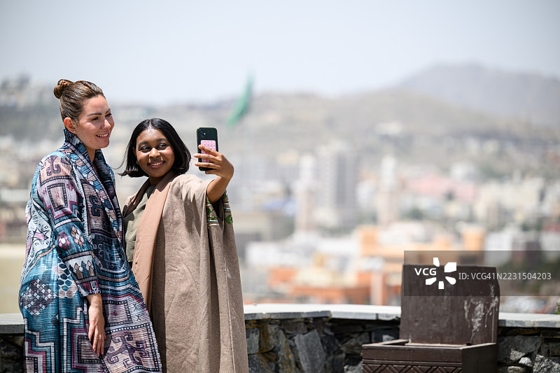 身着阿巴亚服饰的朋友们在沙姆桑奥斯曼城堡的露台上，以阿卜哈城市天际线为背景，开心地自拍图片素材