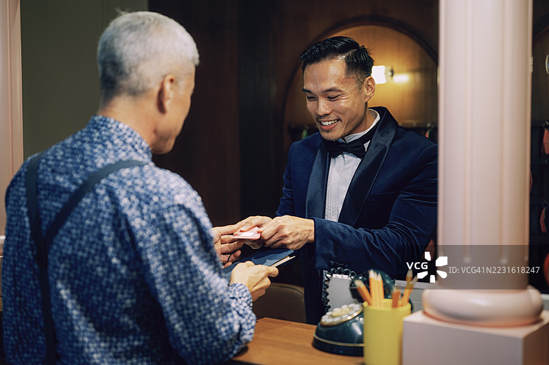 微笑的亚洲男性酒店接待员在前台将房间钥匙递给老年客人图片素材