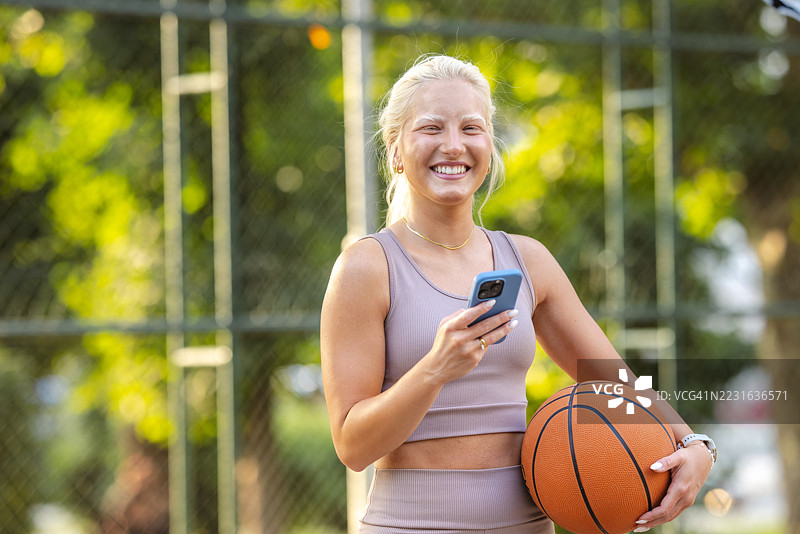 快乐的年轻女性在户外运动场上拿着篮球和智能手机图片素材