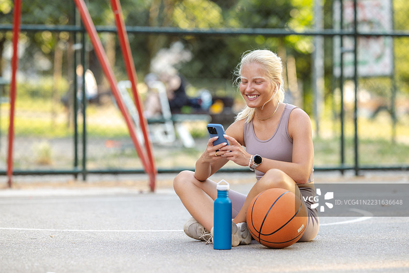 微笑的女性在户外篮球场上放松，手里拿着智能手机和篮球图片素材