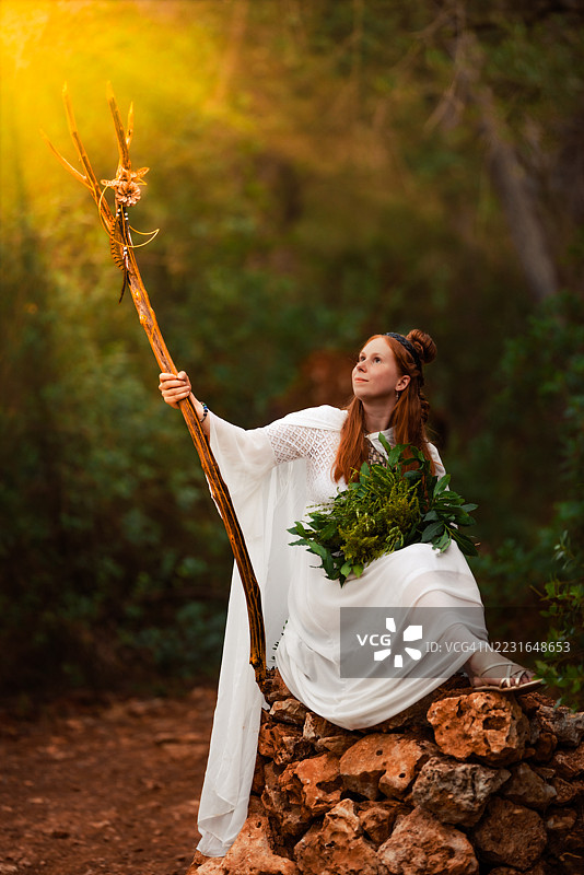 坐在魔法森林石丘上的凯尔特女萨满，手持发光的法杖和草药。图片素材
