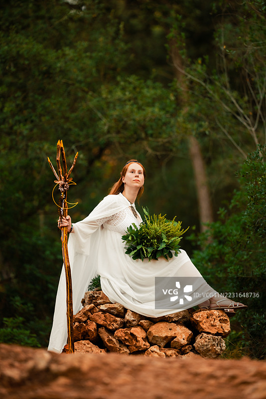 身穿白色长裙的凯尔特女萨满坐在森林山丘上的石头上，手持仪式法杖和草药。图片素材