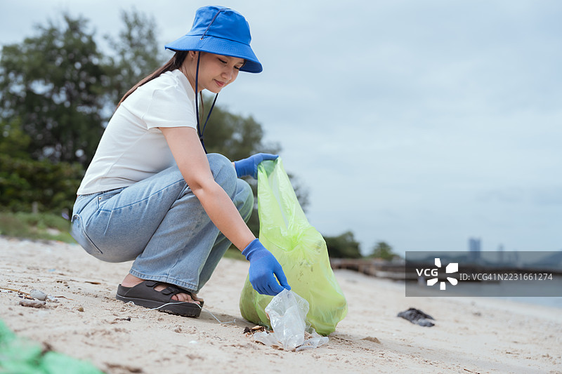年轻女性在阴天戴着手套和蓝色帽子在海滩上捡垃圾图片素材