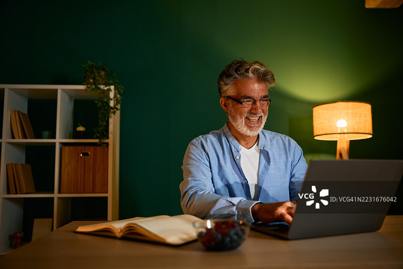 微笑的成熟男性在家中夜晚使用笔记本电脑工作图片素材
