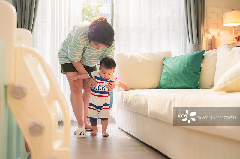 亚洲母亲在金色时光的光线下帮助婴儿男孩迈出第一步，创造了代际之间的联系，通过发展成就。这个真实的育儿内容展示了在关键的童年时刻中给予的关爱指导。图片素材