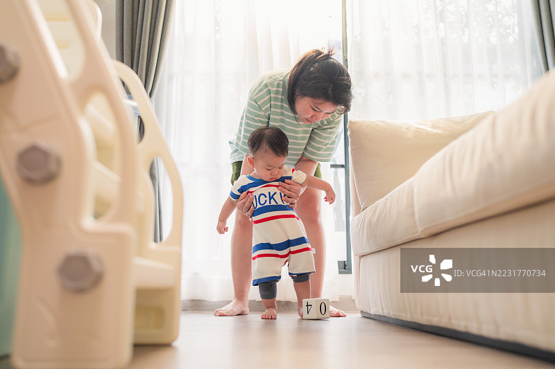 亚洲母亲在金色时光的光线下帮助婴儿男孩迈出第一步，创造了代际之间的联系，通过发展成就。这个真实的育儿内容展示了在关键的童年时刻中给予的关爱指导。图片素材