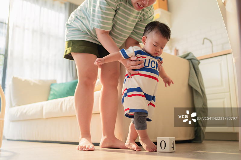 亚洲母亲在金色时光的光线下帮助婴儿男孩迈出第一步，创造了代际之间的联系，通过发展成就。这个真实的育儿内容展示了在关键的童年时刻中给予的关爱指导。图片素材