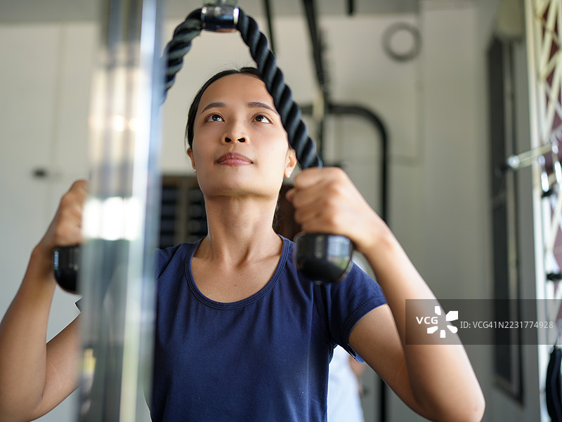 女性在健身房锻炼，专注于使用拉力器进行力量训练。她展现出决心和专注，利用器械提升自己的健身水平。图片素材