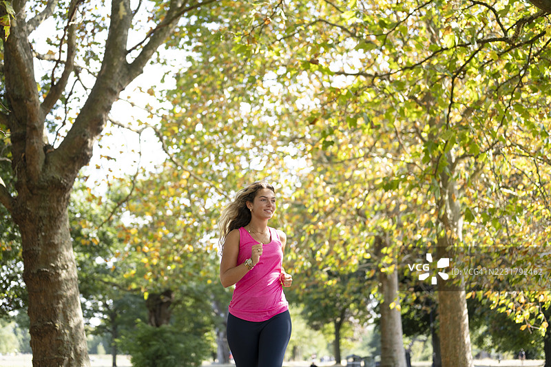 阳光明媚的公园里，一位年轻女子在树林环绕中慢跑图片素材