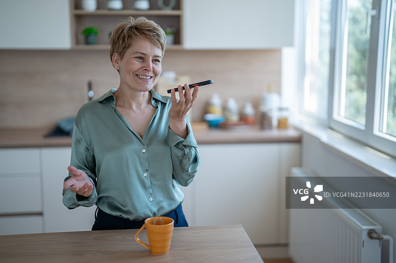 微笑的女性在厨房里用智能手机录制语音信息图片素材