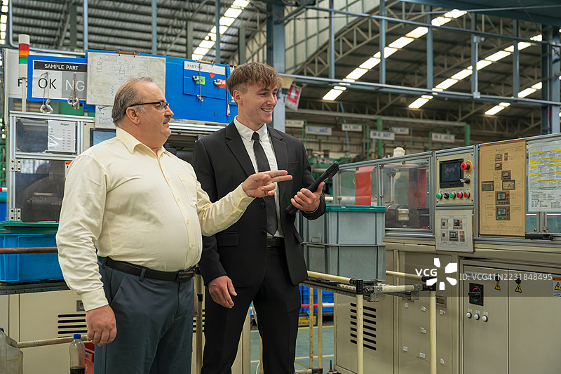 微笑的年长企业主和保险代理人在工厂车间行走，并指向一台机器。图片素材