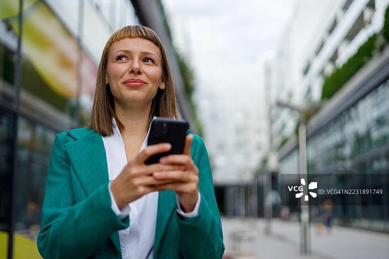 专业女性在时尚的城市摩天大楼间查看智能手机，周围充满城市的活力。图片素材