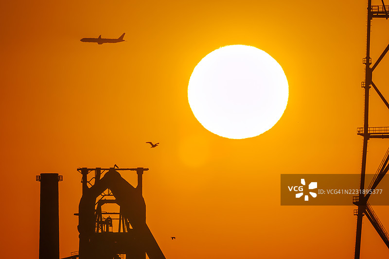 第一缕阳光与一架客机和鸟的剪影。它即将降落在附近的羽田机场。第一缕阳光从运河对岸的烟囱和电厂、钢铁厂上升。神奈川县横滨市，图片素材