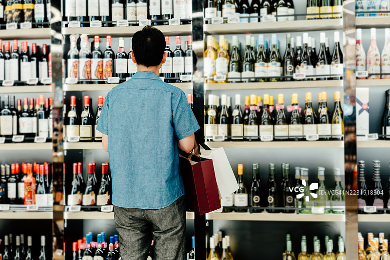 亚洲成熟男性从超市有机便利店的货架上选择一瓶红酒的背影。图片素材