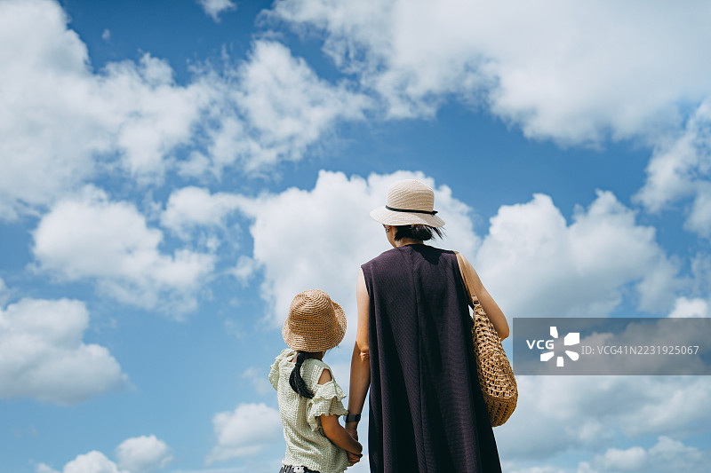年轻亚洲母亲从背后牵着女儿的手，站在美丽的蓝天下，仰望着享受新鲜空气和美景。感受自由，在阳光明媚的日子里放松。母女亲密时刻，家庭的爱与团结。图片素材