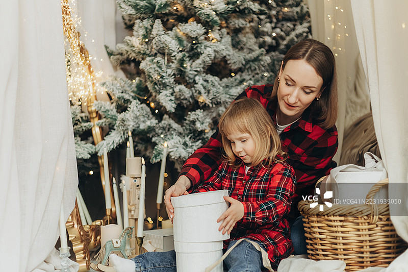 年轻女孩和她的母亲在装饰好的环境中，围绕着雪白的圣诞树打包白色礼品盒。图片素材