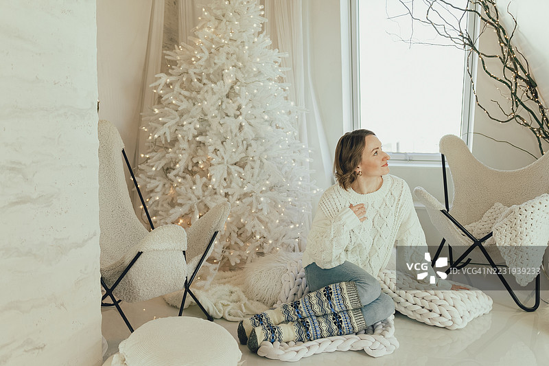 年轻女性在节日的阁楼里，靠在一条厚实的针织毛毯上，旁边是一棵白色的新年树。图片素材