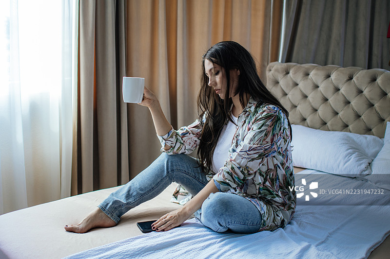 年轻女性在家中床上放松，享受着温暖的咖啡，卧室温馨，清晨的时光。图片素材