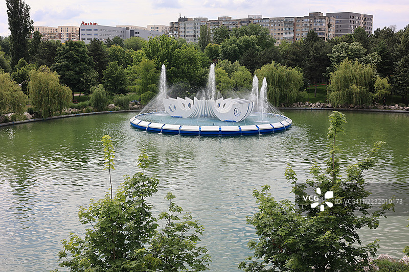 布加勒斯特，罗马尼亚，公园湖中的喷泉，四周环绕着树木和建筑。图片素材
