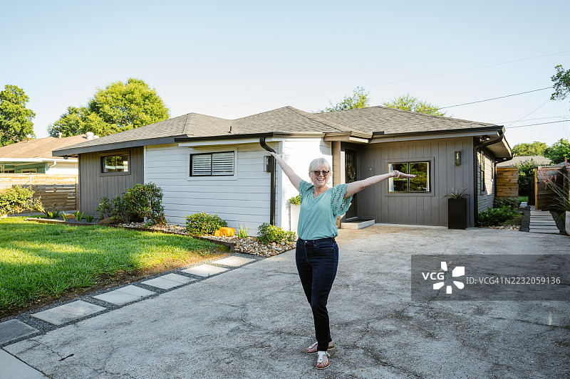 一位老年女性站在她新建的房子前，面带微笑地看向镜头。图片素材