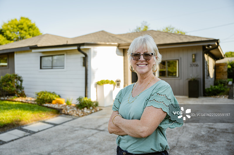 一位老年女性站在她新建的房子前，面带微笑地看着镜头。图片素材