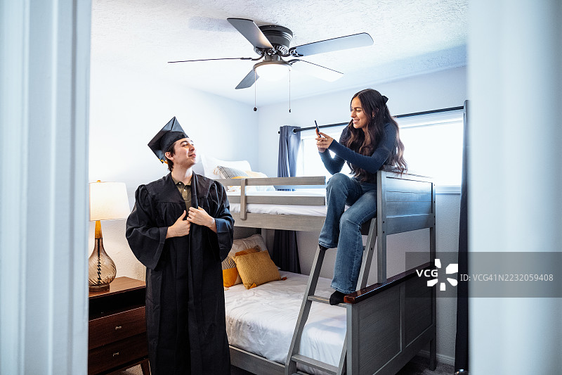 毕业庆祝 - 研究生穿上学位袍和帽子。少女在旁观看哥哥试穿毕业服装。图片素材