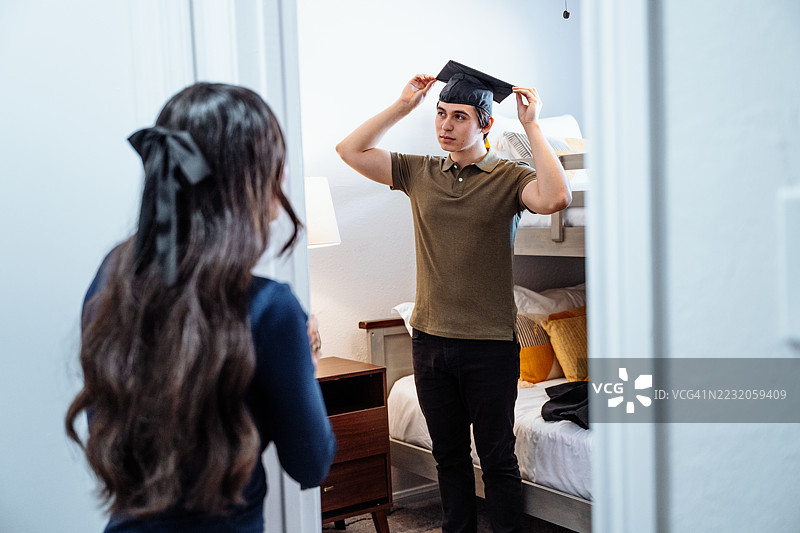 毕业庆祝 - 研究生穿上学位袍和帽子。少女在旁观看哥哥试穿毕业服装。图片素材