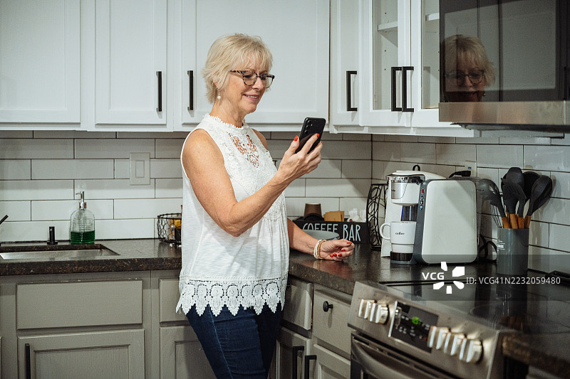 微笑的老年女性在现代厨房中享受早晨咖啡，旁边的台面上摆放着新鲜的早餐食材，正在看手机。图片素材
