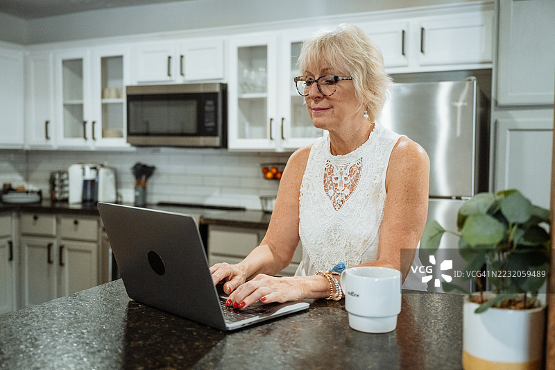 微笑的老年女性在现代厨房的台面旁享受早晨咖啡，同时使用笔记本电脑。图片素材