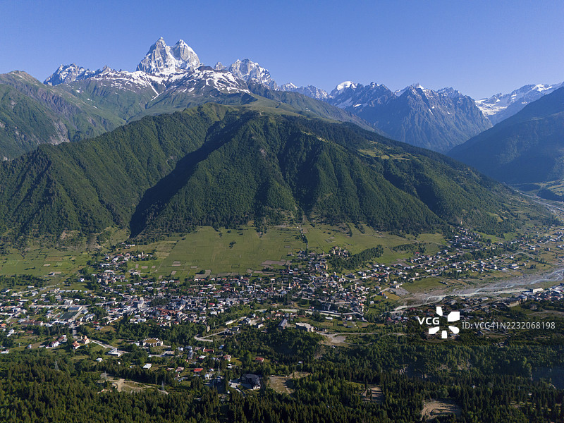 从空中俯瞰，绿色山谷中的村庄被高耸的、白雪皑皑的山峰环绕，天空湛蓝，这里是格鲁吉亚斯瓦涅季高加索山脉的梅斯蒂亚，穆尔赫拉河畔的乌什巴山。图片素材