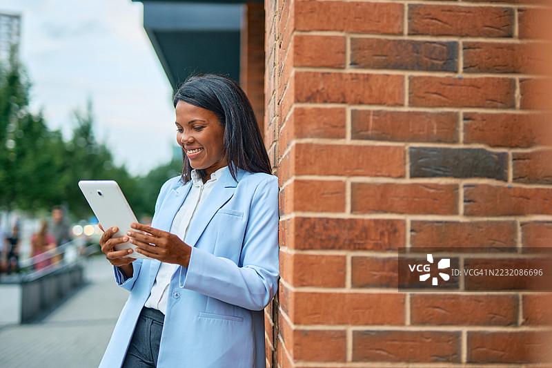 微笑的女商人在城市的砖墙旁使用平板电脑图片素材